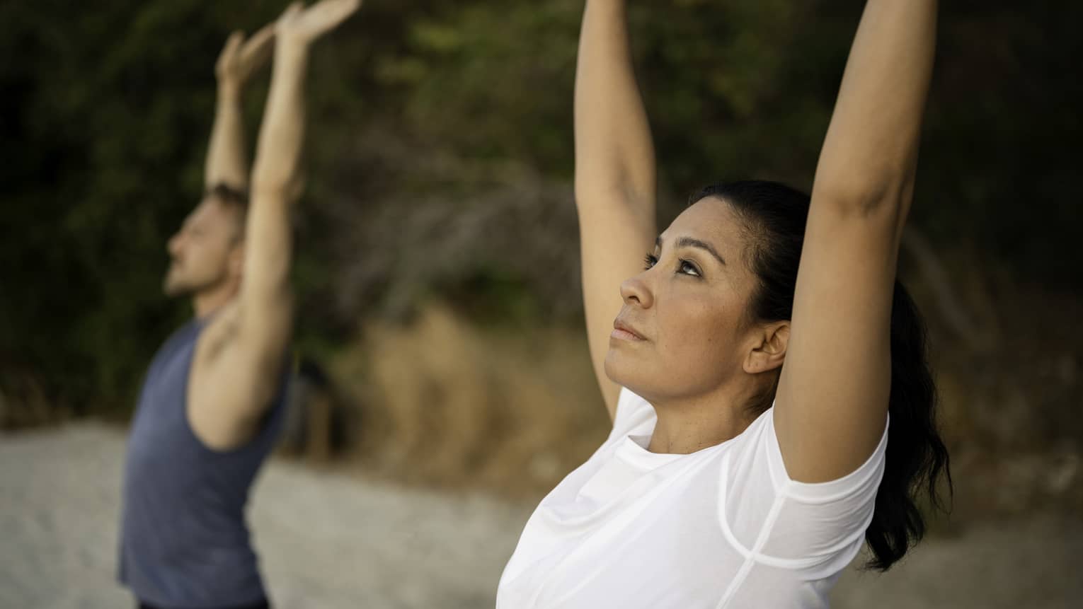 A woman in a white t-shirt and a man in a blue muscle tee stand on the beach stretching their arms above their heads