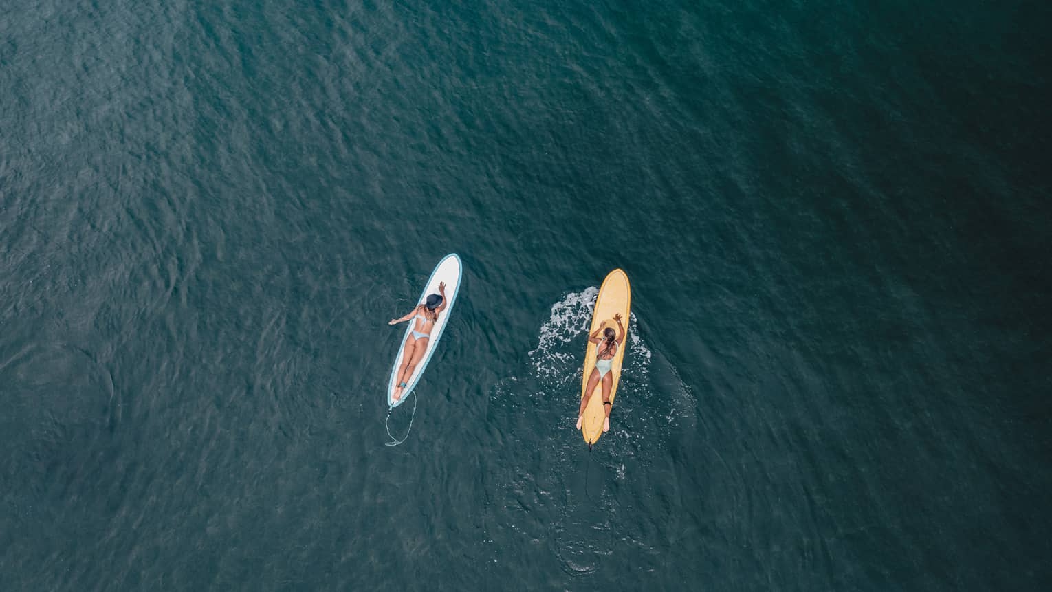 Aerial view of two guests lying on their stomachs on surfboards facing each other on calm water, each wearing a safety leash.