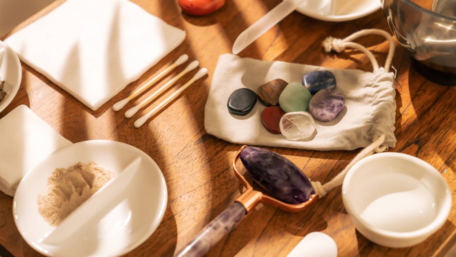 Face roller, gemstones, cotton swabs and a variety of small white bowls and other instruments sit on a sun-lit wooden table