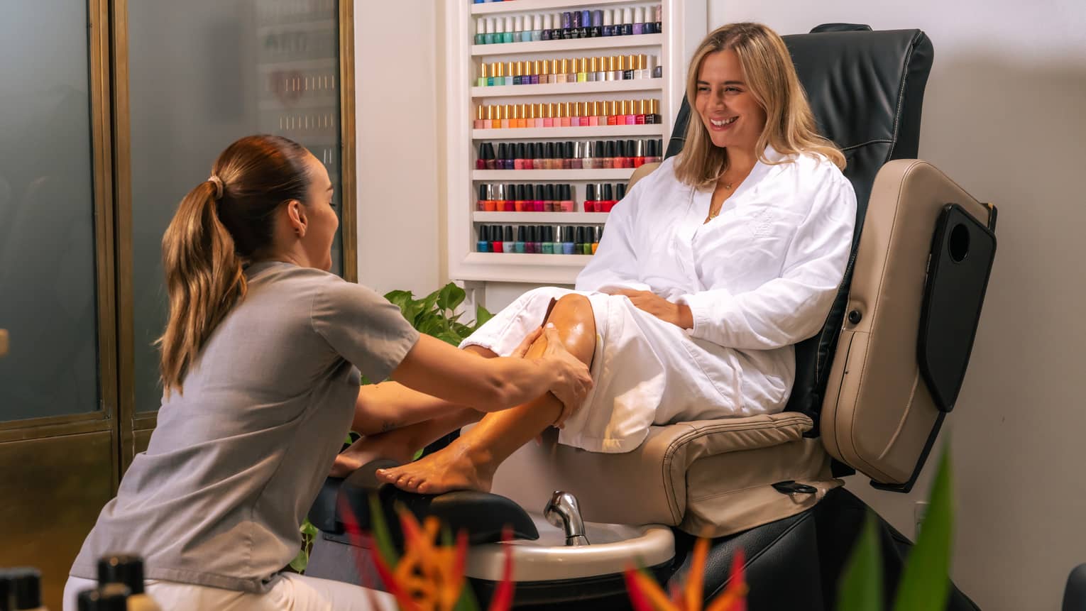 Person with shoulder-length blonde hair and wearing a white spa robe sits at a pedicure station as an attendant with light-brown hair tied up in a ponytail and wearing a grey polo shirt with white pants washes the person's shins