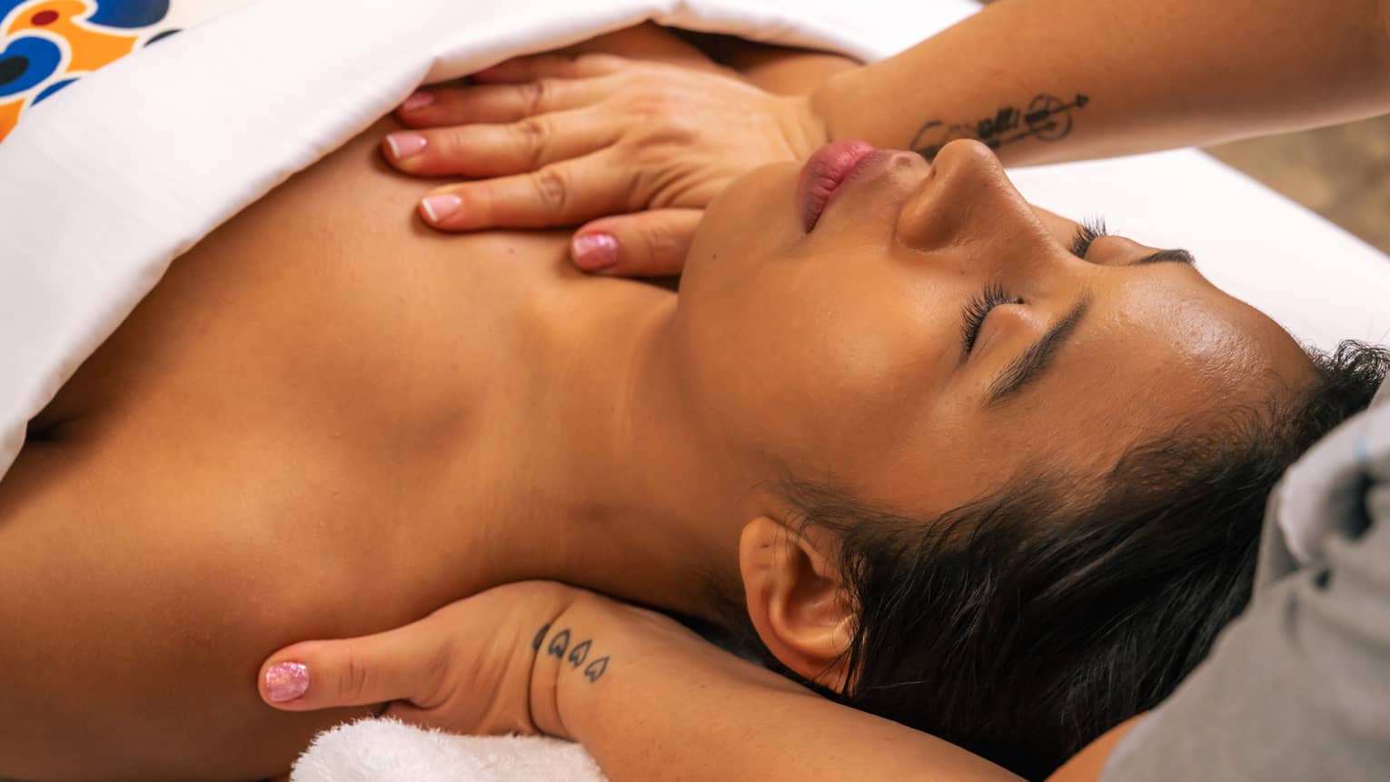 Person lies on their back beneath a white blanket with their eyes closed as a pair of hands massages the back of their shoulder and applies pressure on sternum