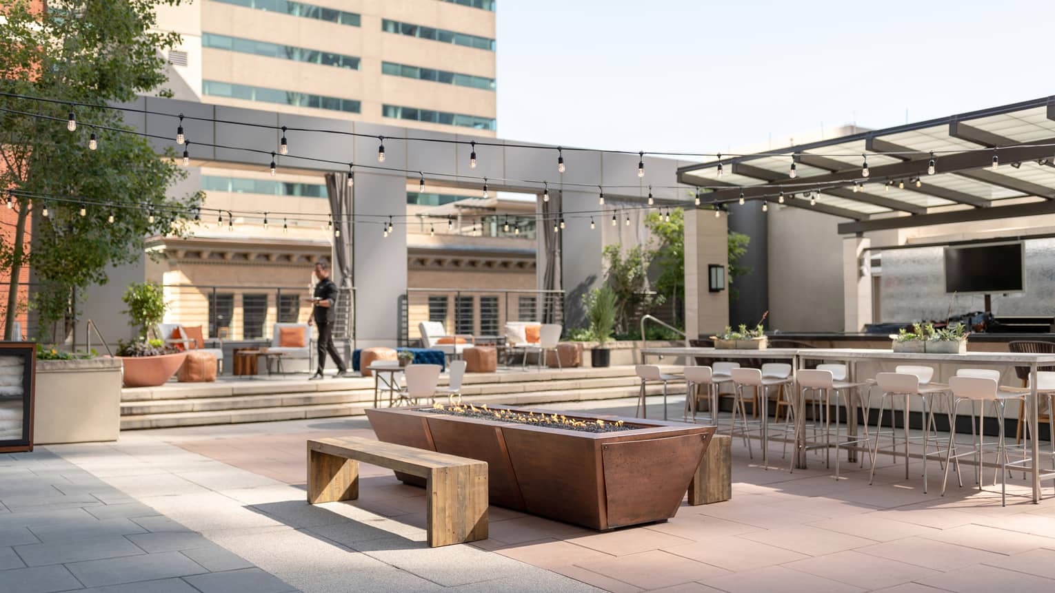 A staff member walking across a pool courtyard outside, a fire pit, pool, and seating can be seen.