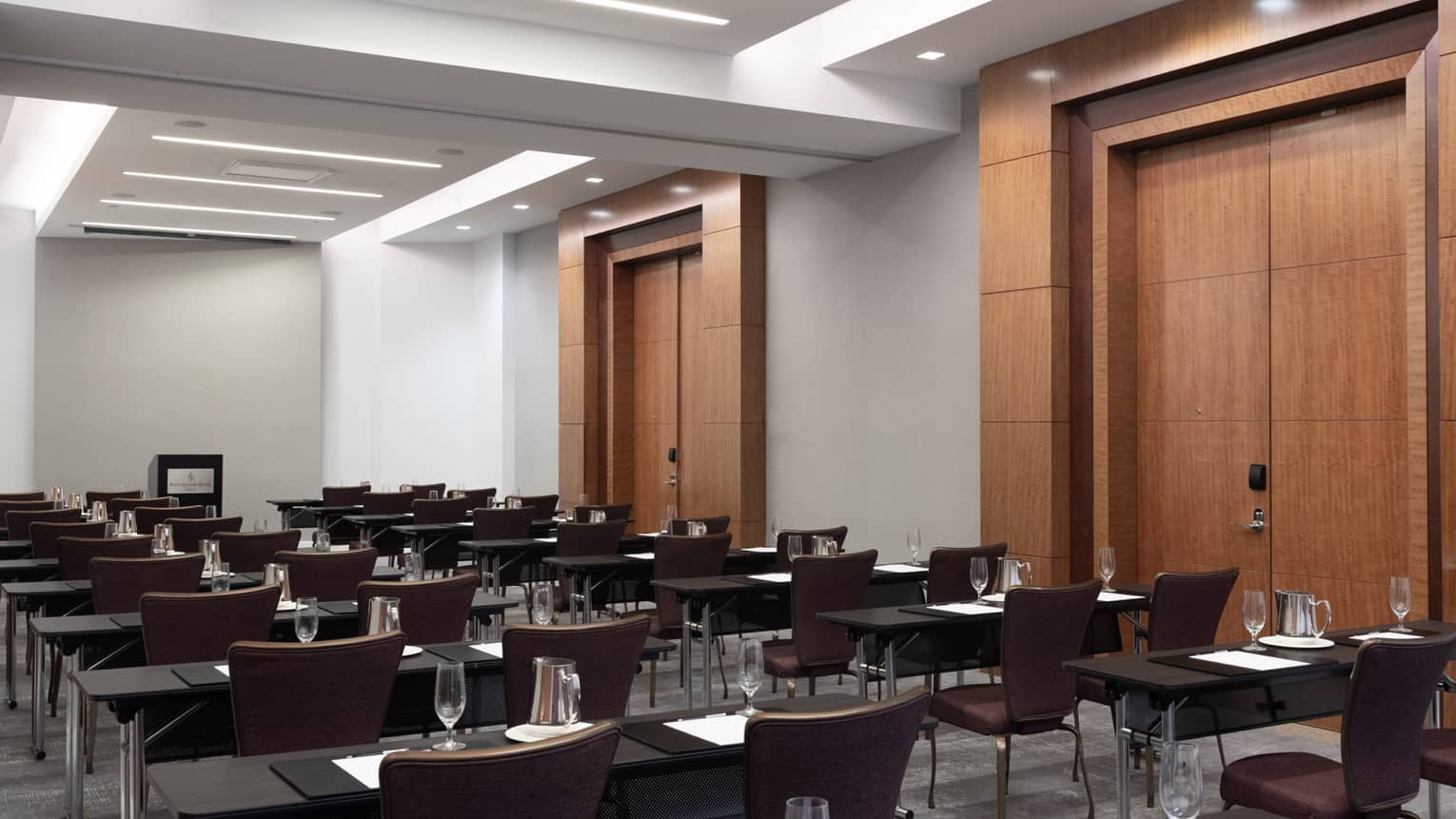 Rows of desks and brown chairs, with water glasses on each desk, in a long room facing a podium.
