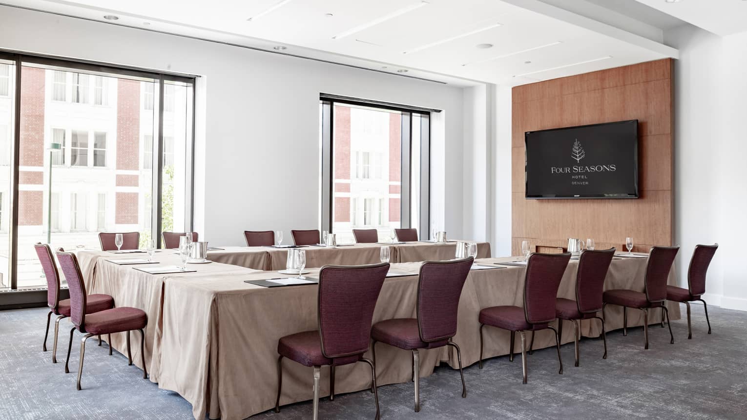 A "U" shaped table covered in a beige tablecloth with brown chairs around it, in a room with large windows on one wall.