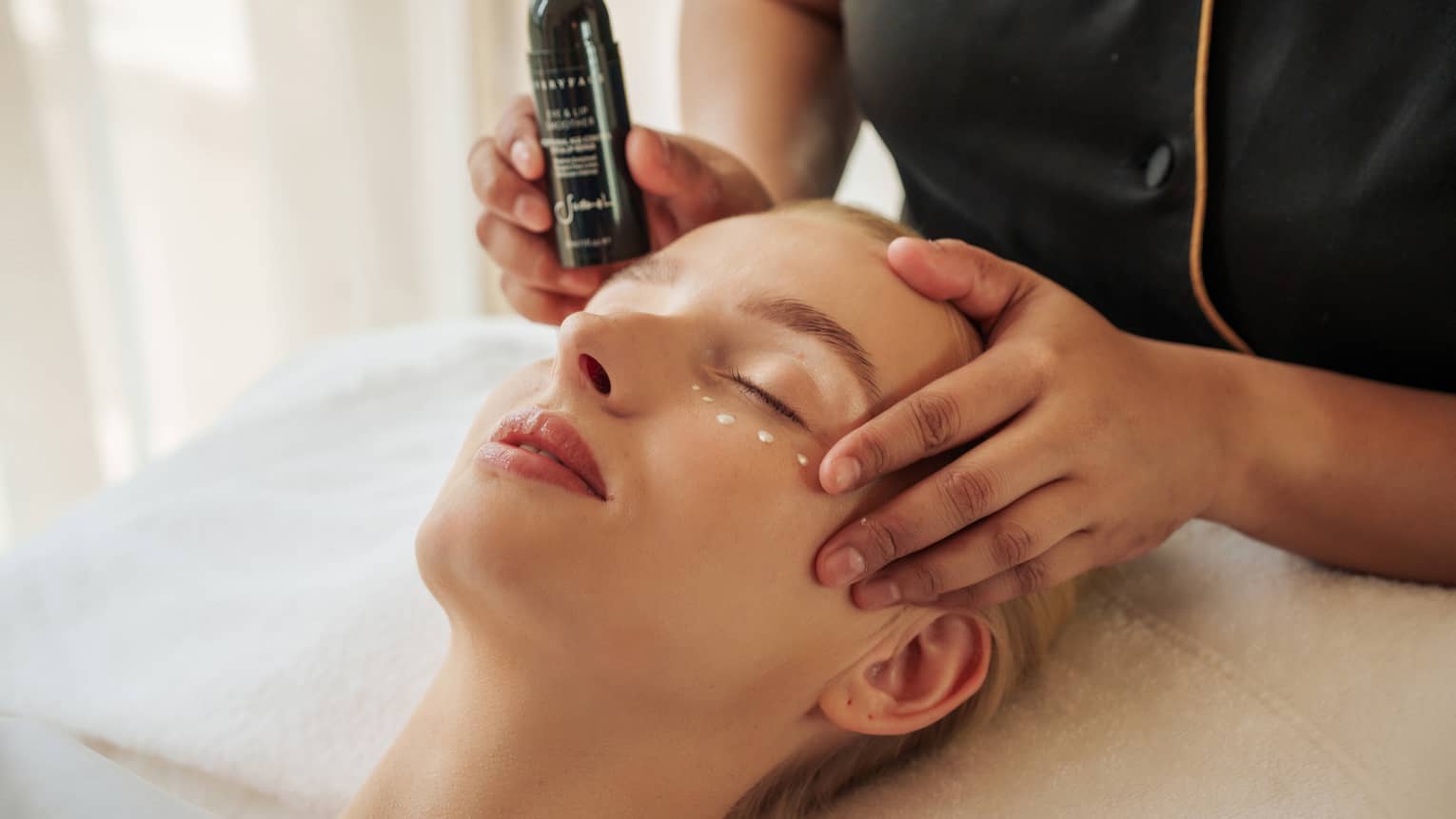 Guest lies with eyes closed as a spa attendant places product under their eyes