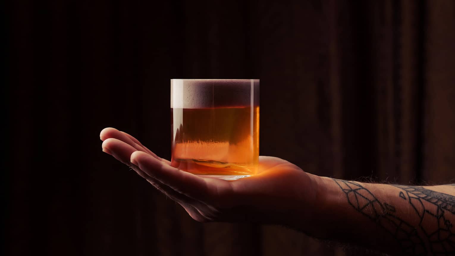 A tattooed arm reaches in from outside the frame and holds a rocks glass filled with an orange-colored cocktail on his outstretched hand
