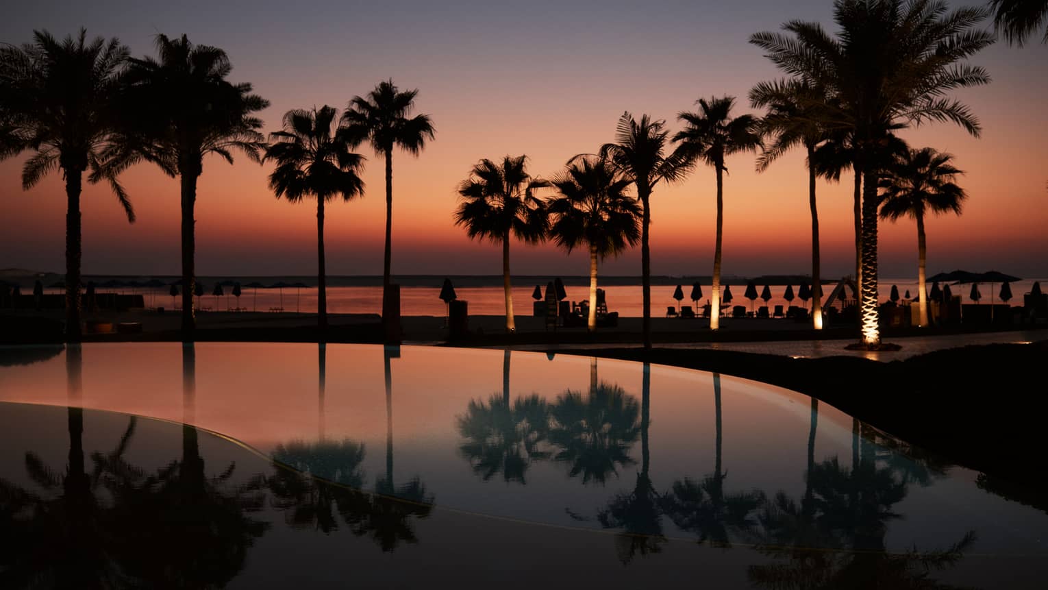 A palm tree?lined pool with the beach in the background all lit under a dark orange and blue sky as the sun sets