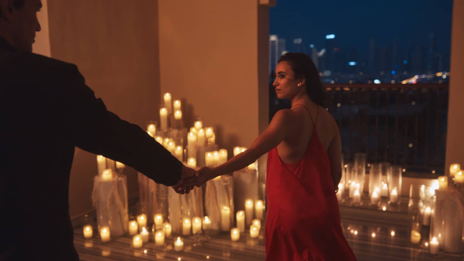 Person wearing a red dress leads another person who is silhouetted in the shadow by the hand toward a terrace filled with lit candles at night