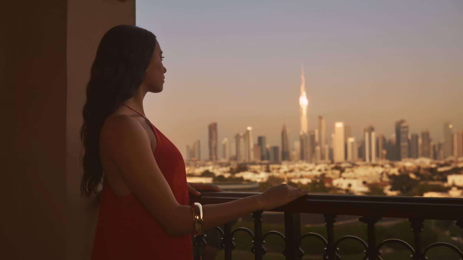 Person with long brown hair standing on balcony with hand resting on railing, looking out to a city at dusk
