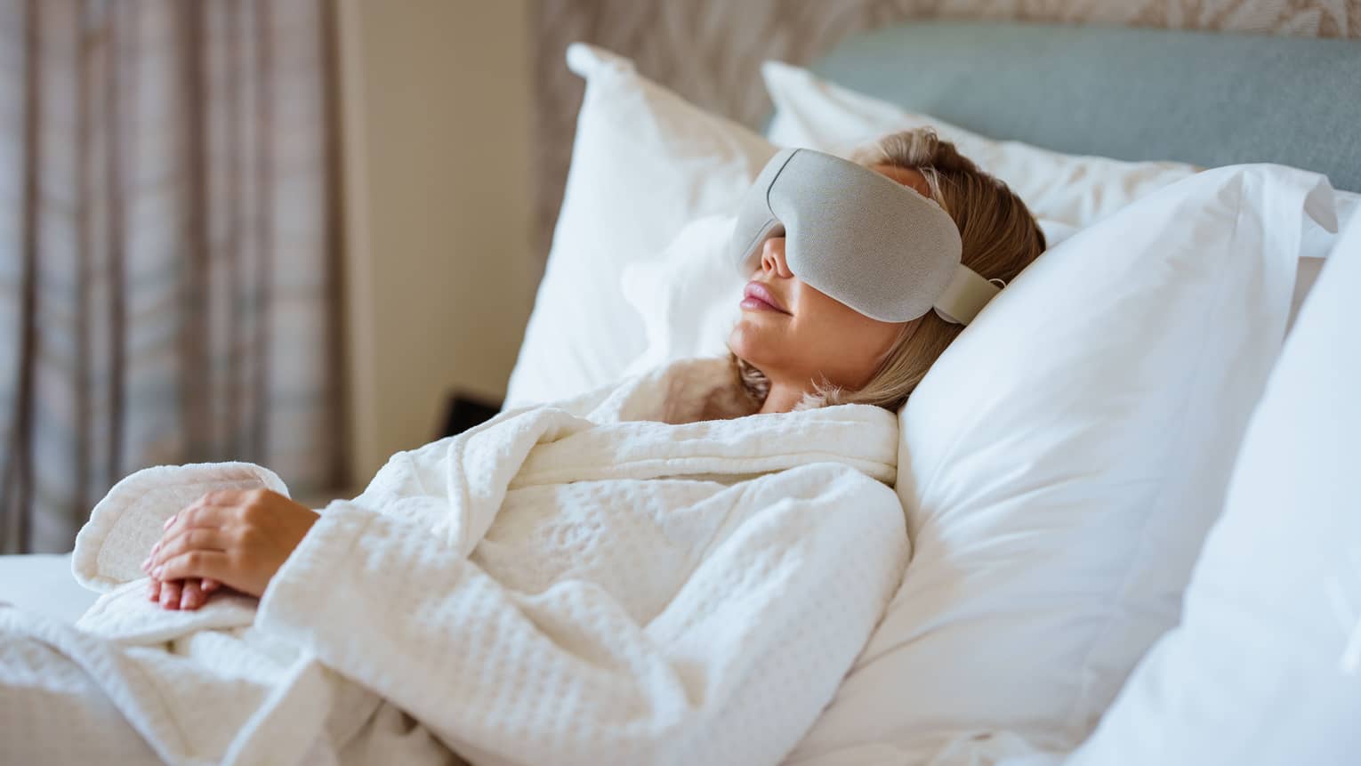 Person wearing white spa robe lays back on a bed with large white pillows wearing a high-tech eye mask