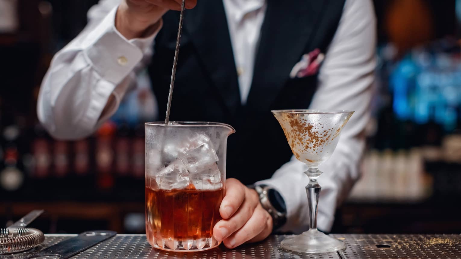 Close-up of bartender stirring liquor over ice in glass pitcher next to cocktail glass on bar 
