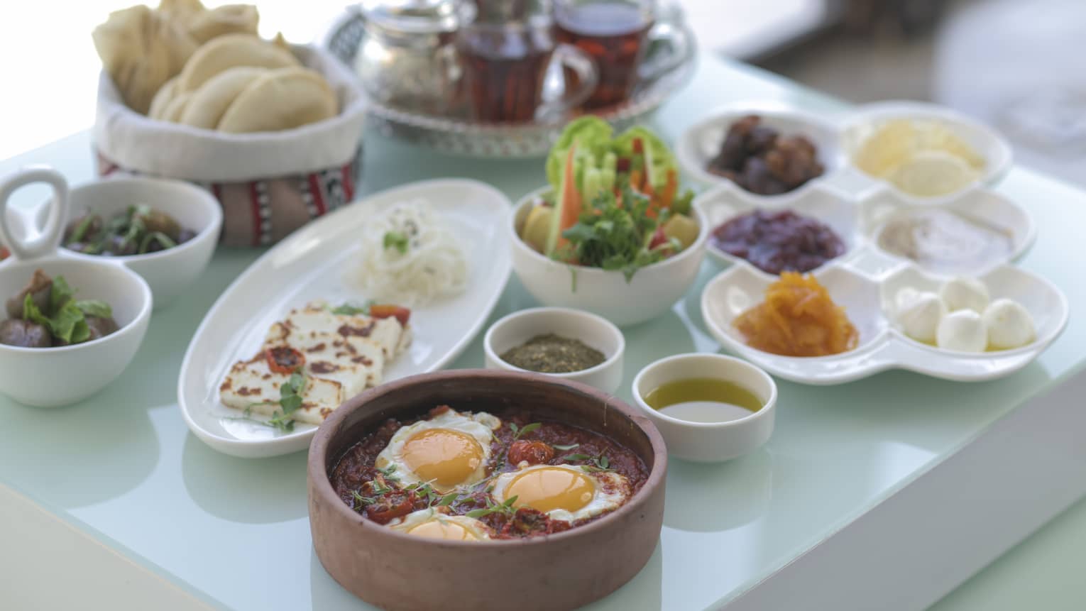 An assortment of traditional Arabic breakfast food on white platters, three eggs in a clay pot, pita in a basket and a glass tea tray