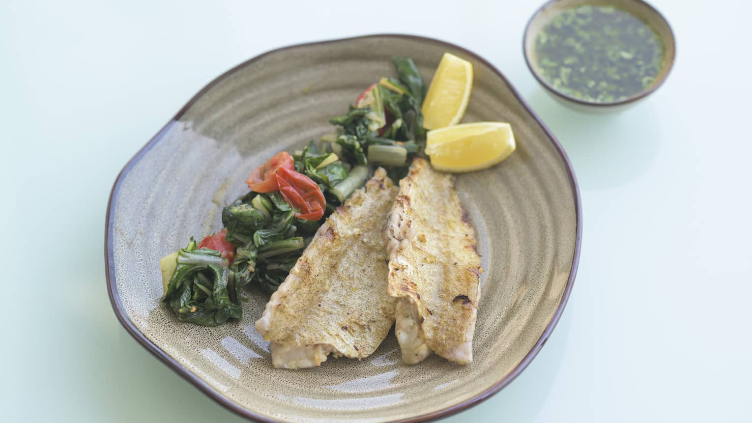 Two fillets of grilled Hamour fish arranged on a round tan plate along with a row of greens, tomatoes and two lemon wedges