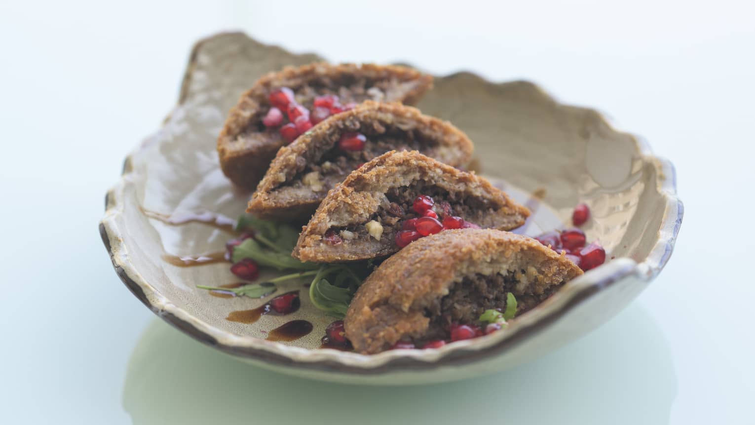 Cream-colored leaf-shaped platter filled with four pieces of Kibbeh Sajyeh on a bed of lettuce, sprinkled with pomegranate seeds
