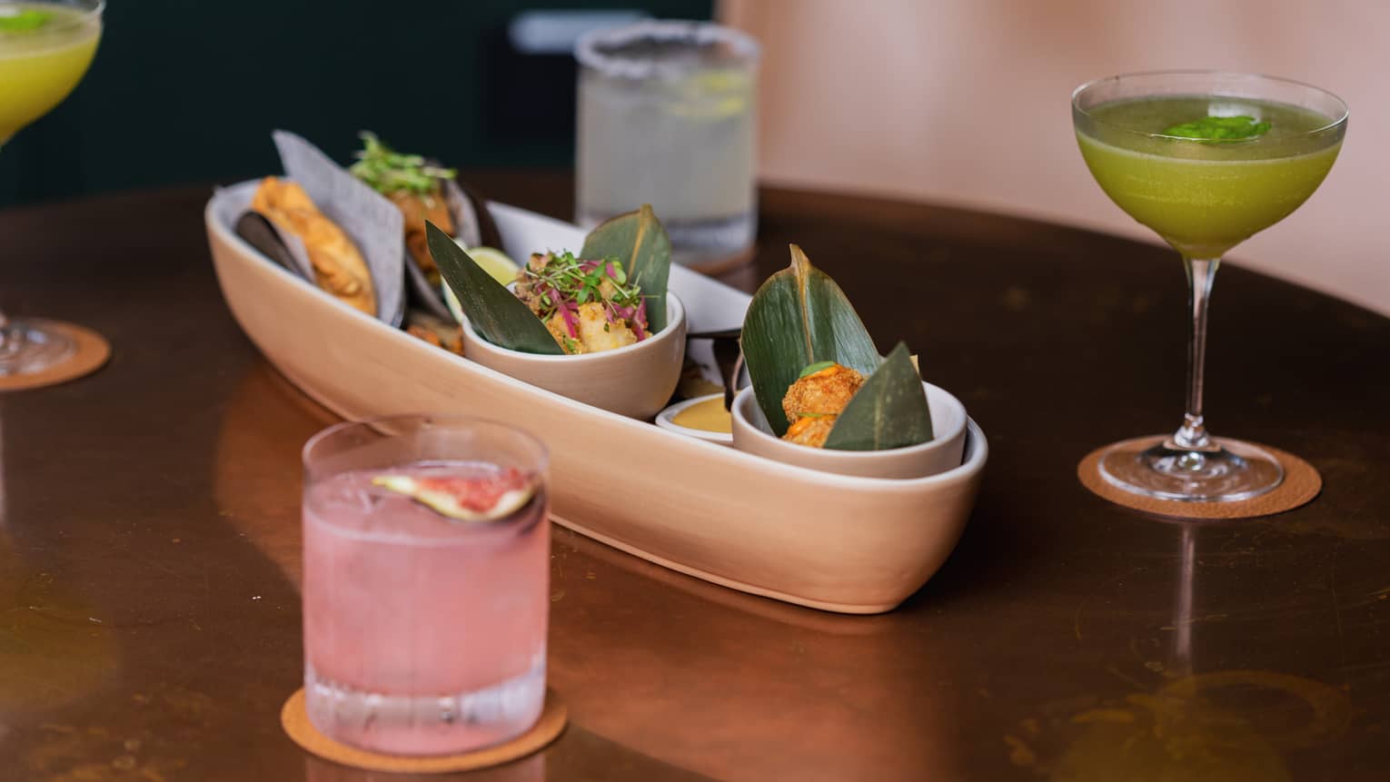 A tan oblong platter holding three small round bowls of food sits in the middle of four cocktails on a table