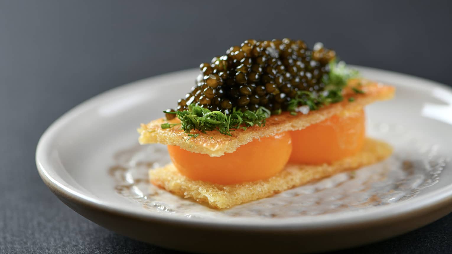 Two toasted egg yolks topped with caviar and herbs on a white round dish set on a black table