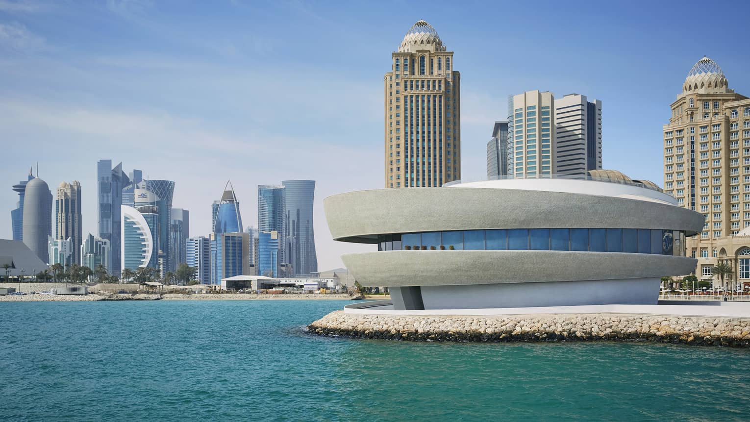 Exterior view of Nobu Doha, a round building with windows in the center sitting on the edge of the water with the skyline behind it
