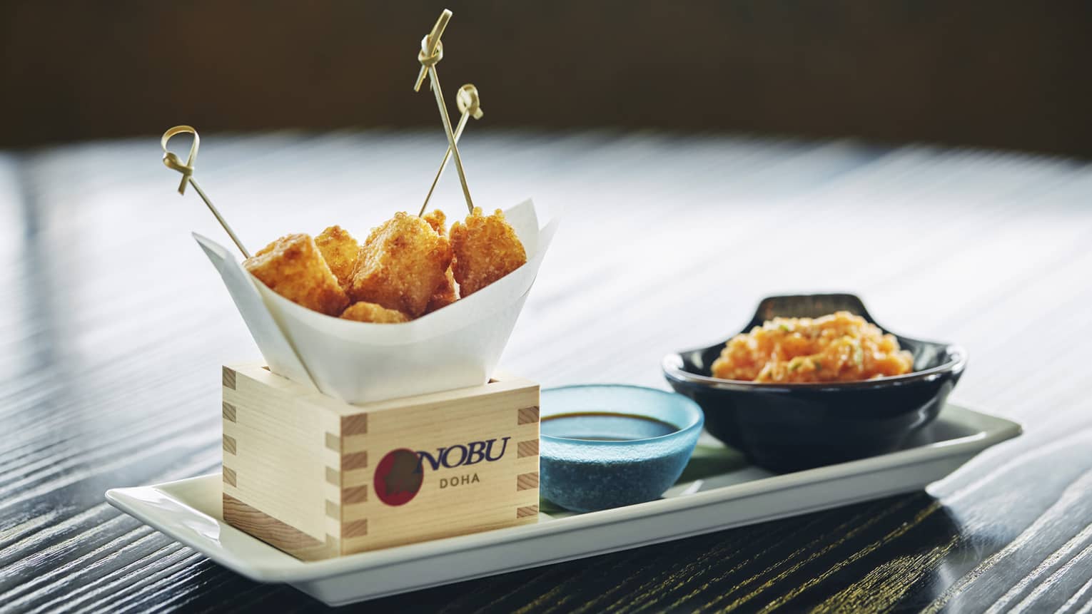Close-up of white platter, wood box with Nobu logo, fried appetizers on toothpicks, dish with sauce