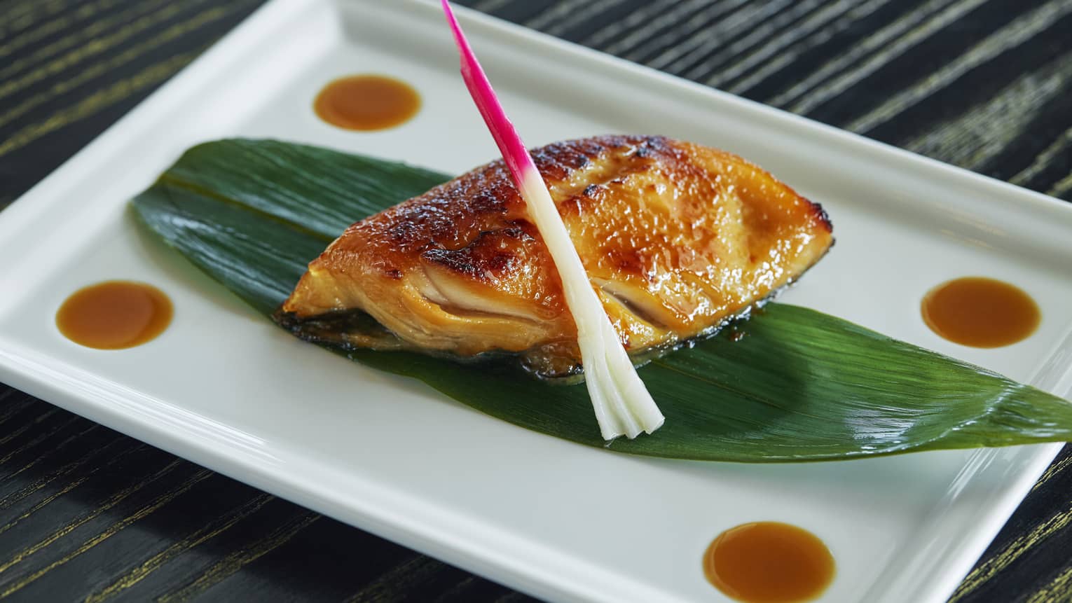 Black Cod Yuzu Miso fish fillet marinated, oven-baked on grilled seafood filet on leaf on white platter
