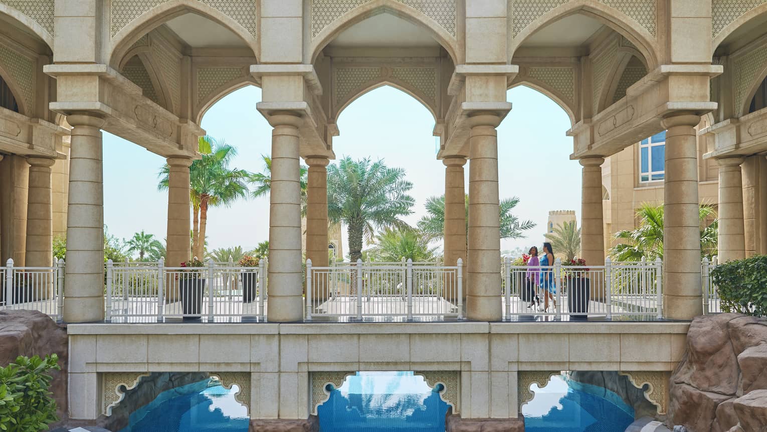 Two women walk across outdoor covered pathway with arches, pillars over blue water 