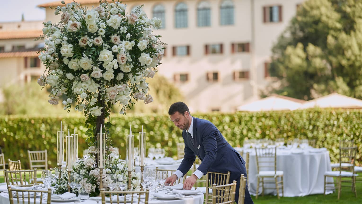 Man in suit sets garden wedding reception tables with large white topiary and hotel in backdrop