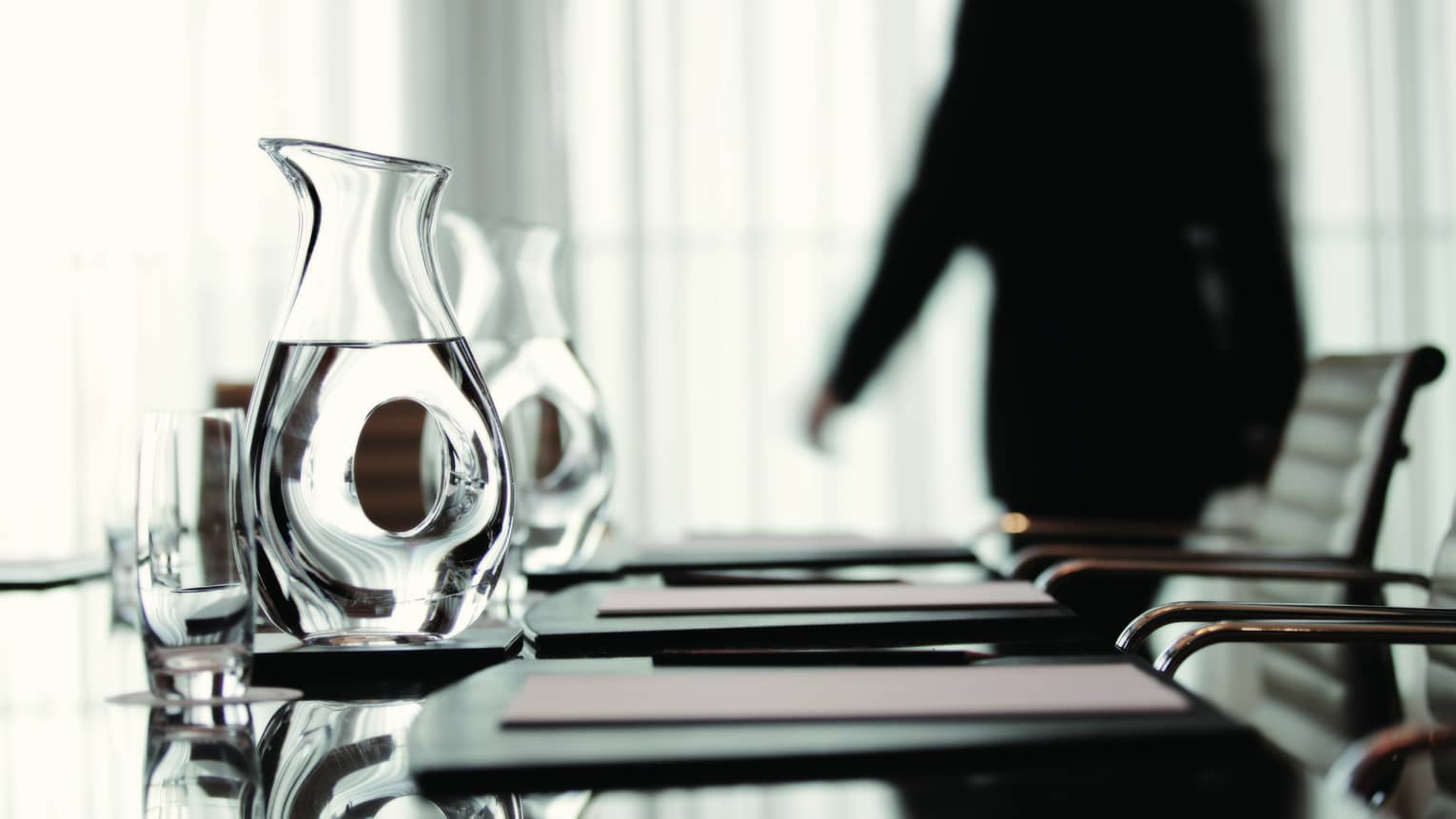 Person wearing business suit walks behind meeting table with agendas, glass water jugs