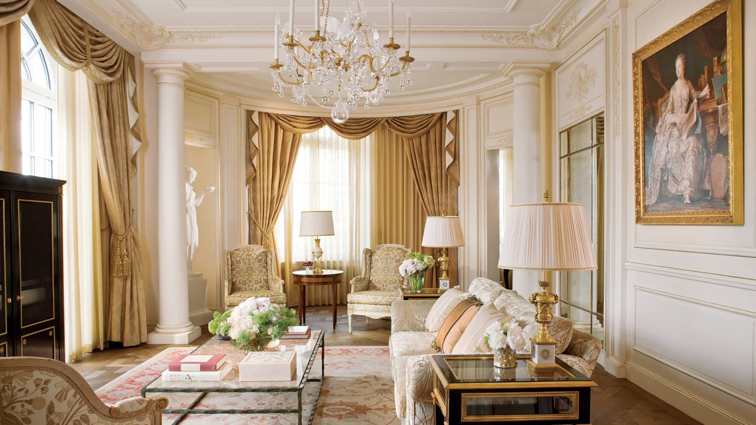 Luxury sitting room with gold accents, ornate drapery, armchairs, chandelier and floral centrepiece on a coffee table, framed by grand columns and a statue