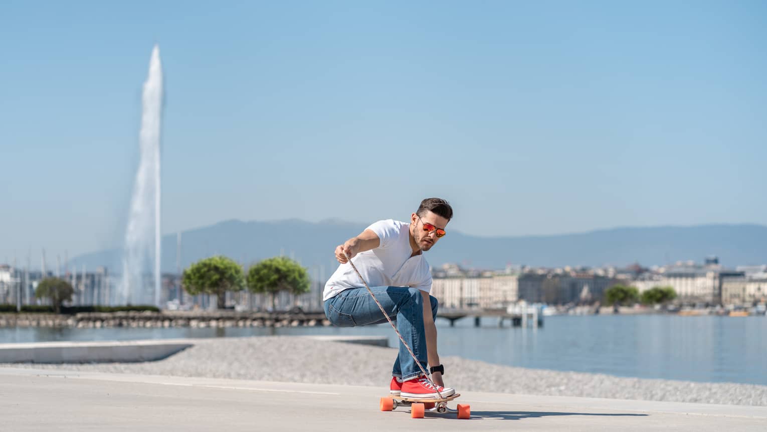 Man longboarding on sidewalk along Lake Geneva with Jet d'Eau fountain in backdrop