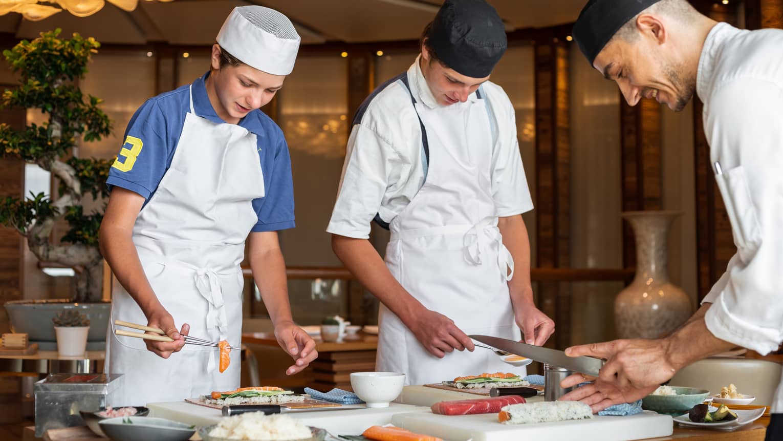 Two teenagers wearing aprons and chef hats learning to make sushi with a sushi chef
