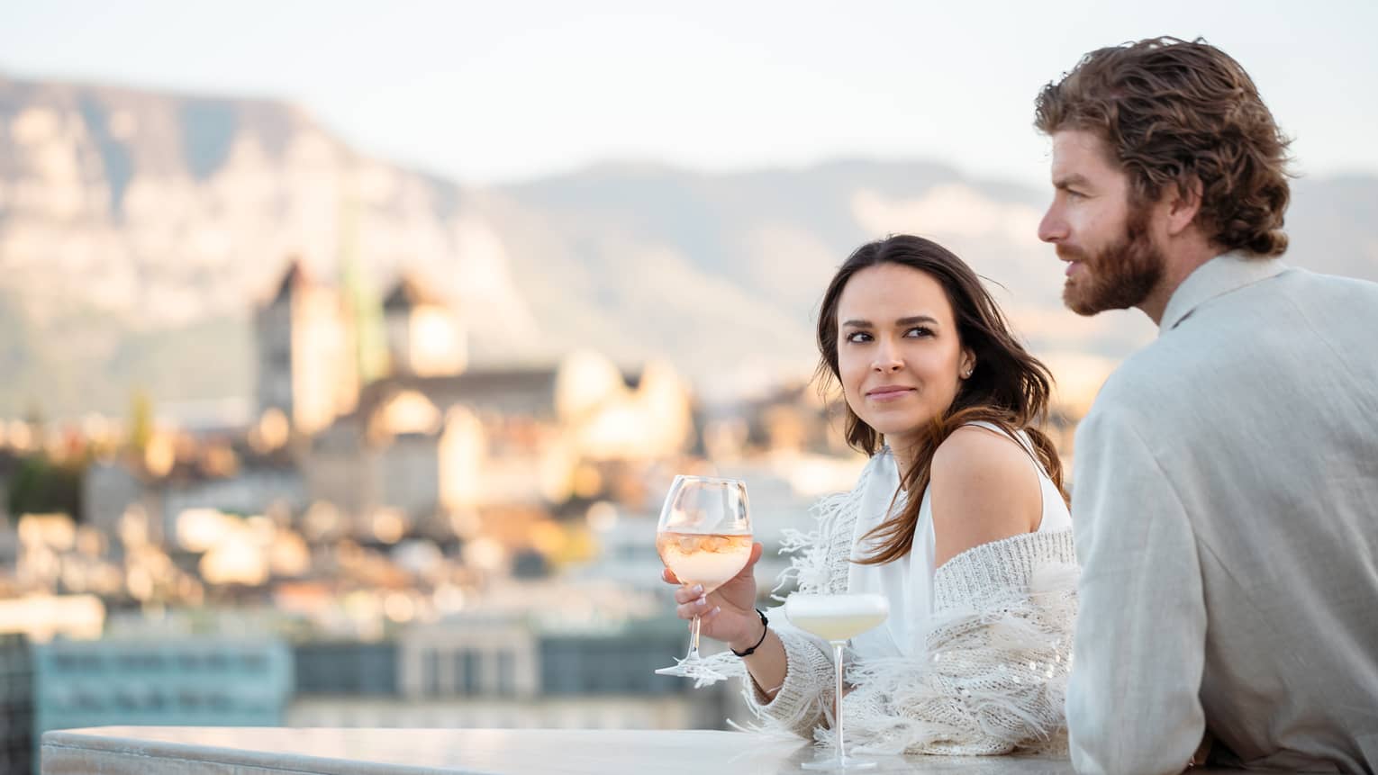 Couple leaning on terrace bar, with wine in hand, overlooking Geneva