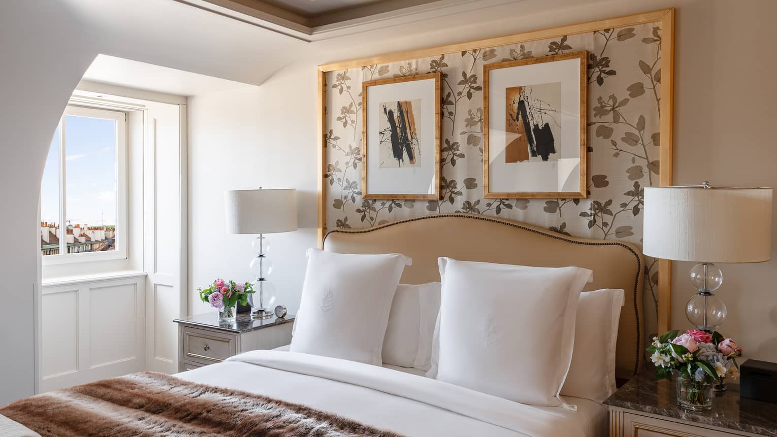 Luxury hotel room with a king bed, marble-topped bedside tables with lamps and flowers, floral wallpaper, framed artwork and a window with city view