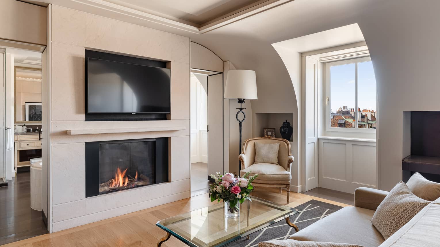 Luxury hotel room with a modern fireplace, wall-mounted TV, beige sofa, armchair, glass coffee table with flowers and windows offering city view