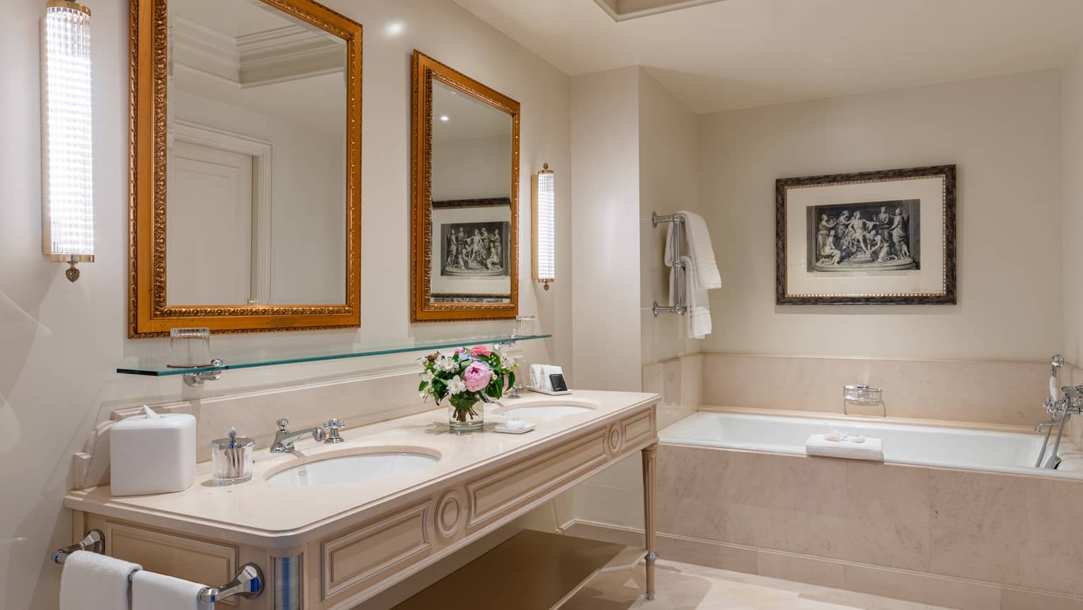 Luxury hotel bathroom with a double sink vanity, gold-framed mirrors, wall sconces and a bathtub framed by elegant artwork