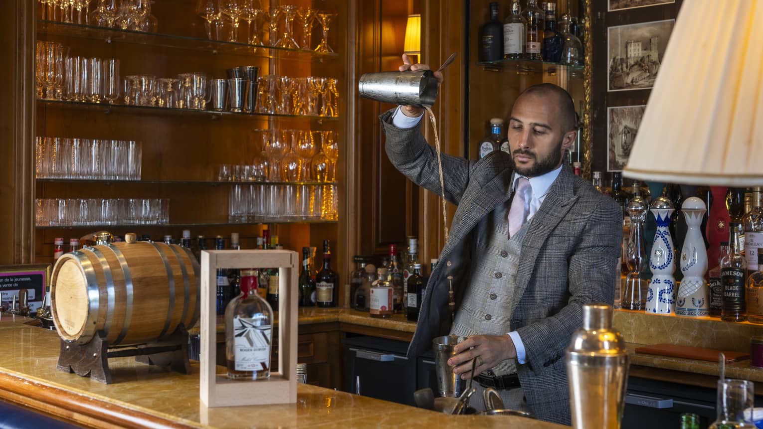 A bartender mixes a cocktail with a shaker behind a well-stocked bar.