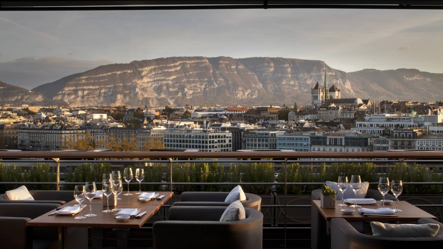 Outdoor patio with armchairs, tables set with wine glasses, overlooking city and mountain on sunny day