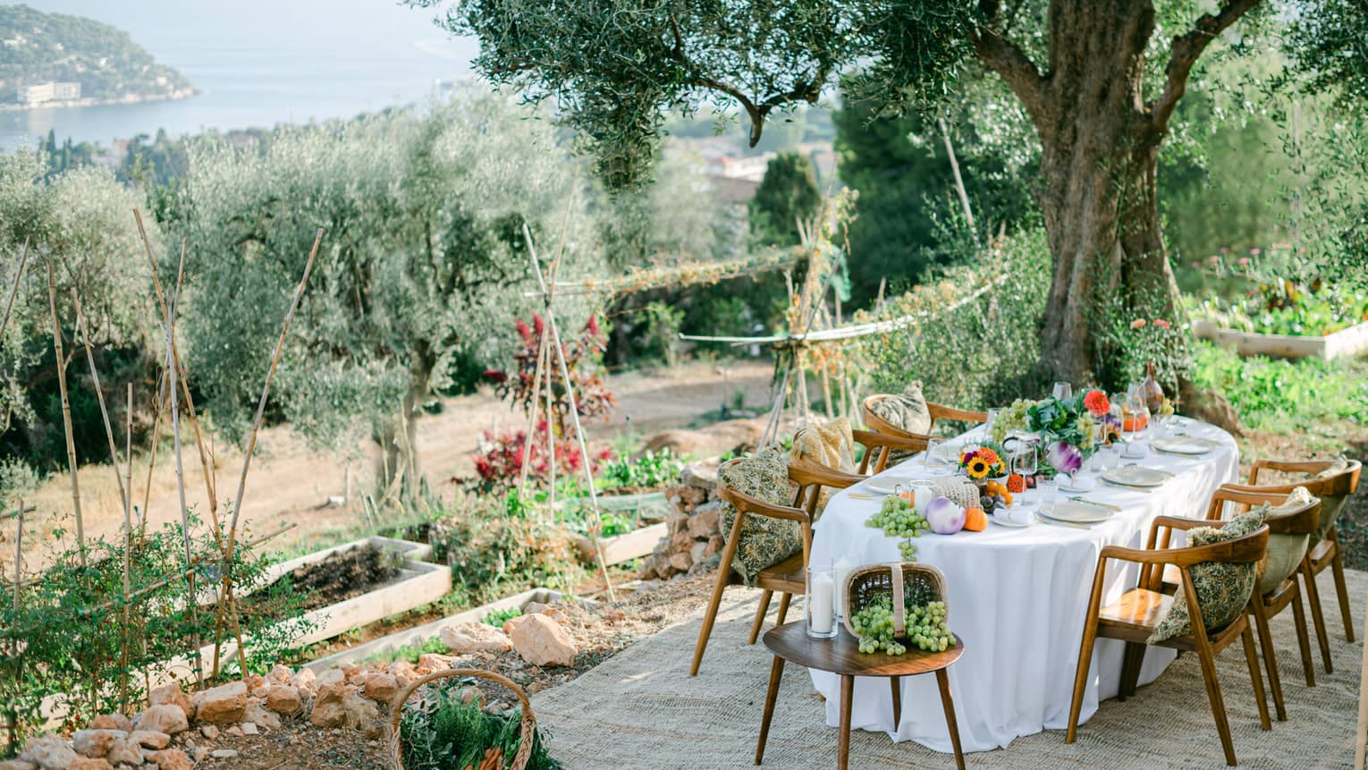 Garden dining setup with oval table draped in white and garden bounty down centre, and six chairs with pillows