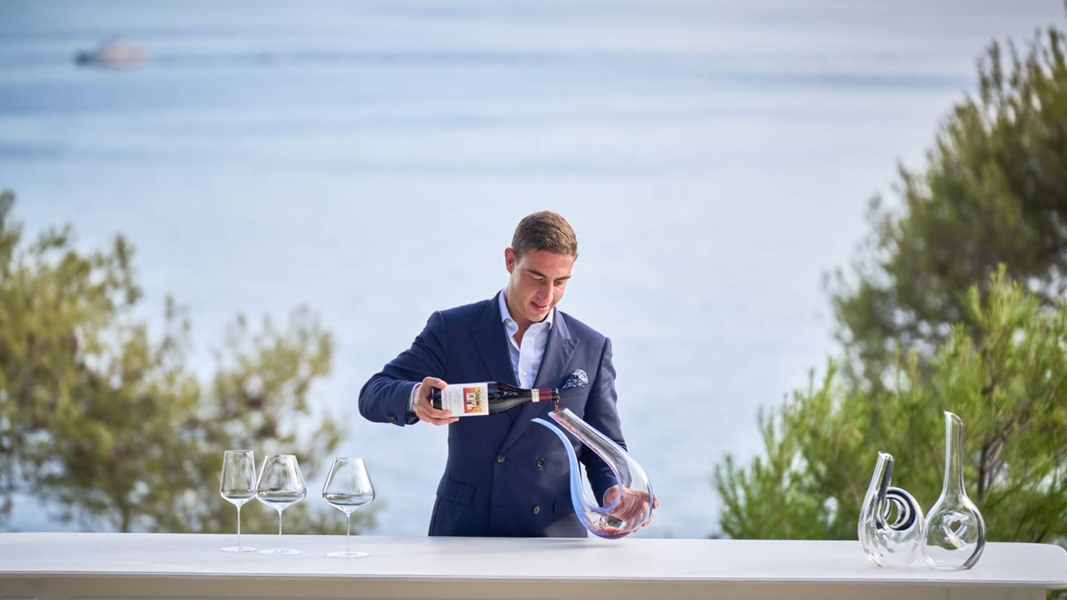 Head Sommelier Alessandro Nigro Imperiale stands behind waterfront table, decanting a bottle of red wine 