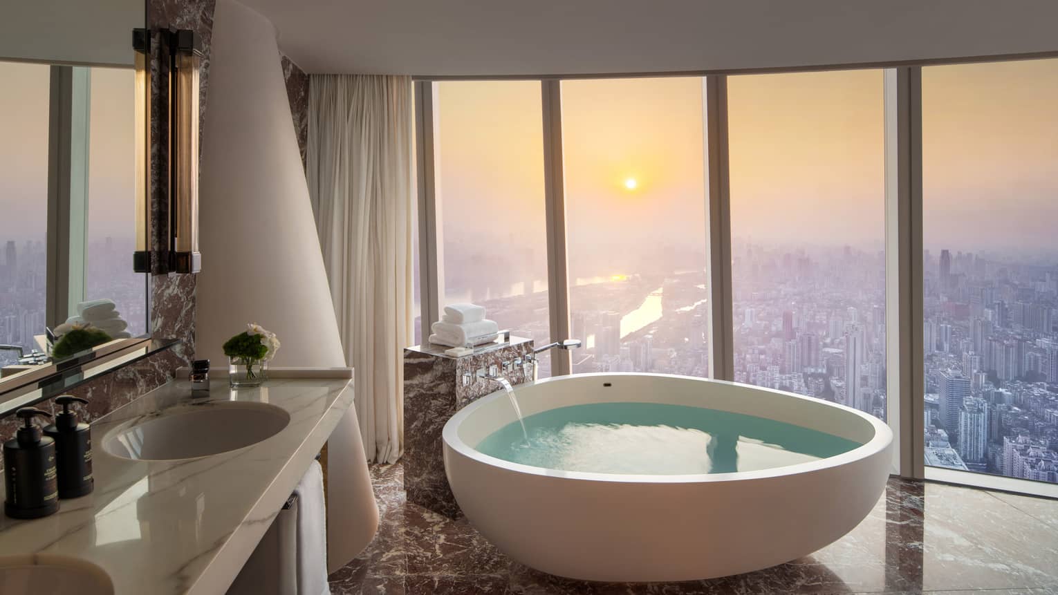 Large bathroom of a hotel room, featuring a large standalone tub next to floor-to-ceiling windows looking out to cityscape
