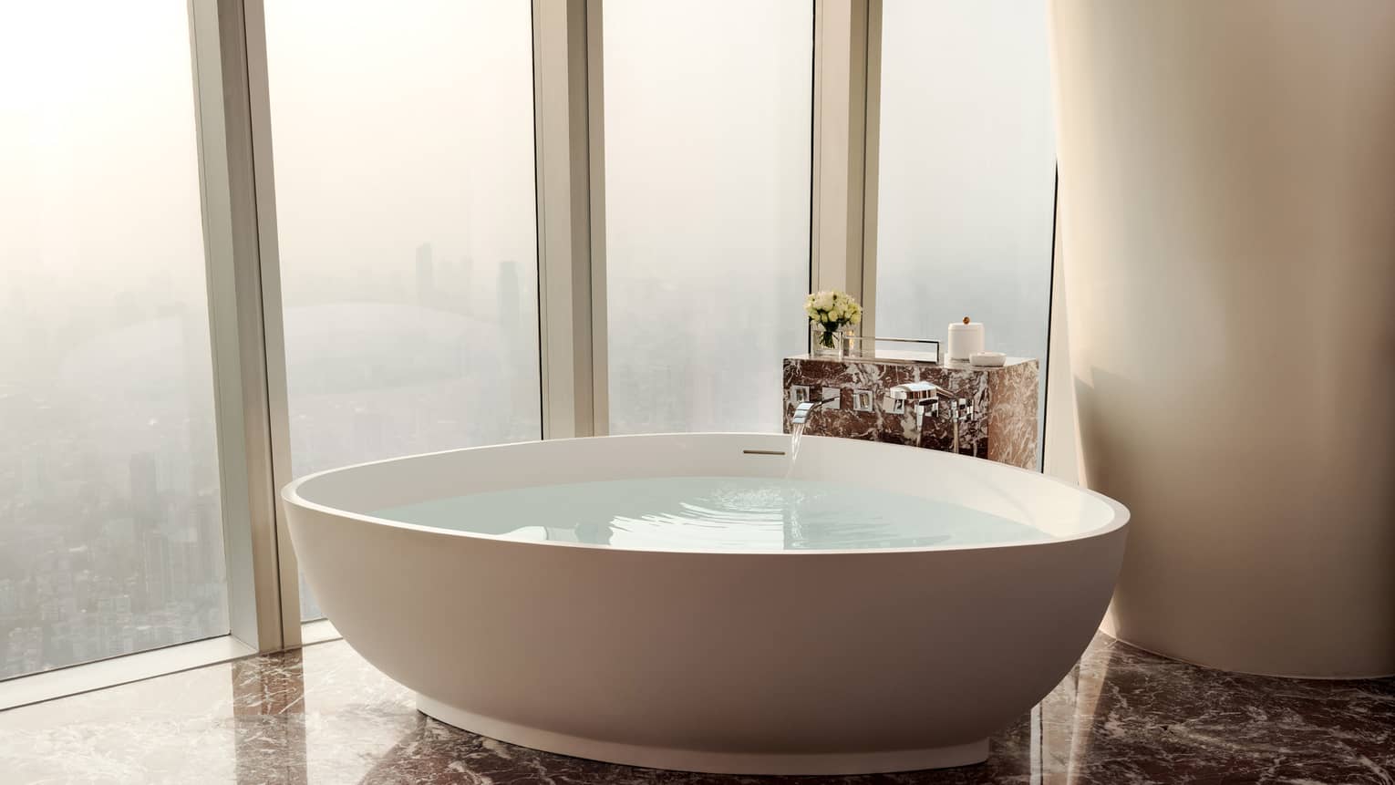 Modern bathroom of hotel room, featuring a large standalone tub next to floor-to-ceiling windows looking out to city