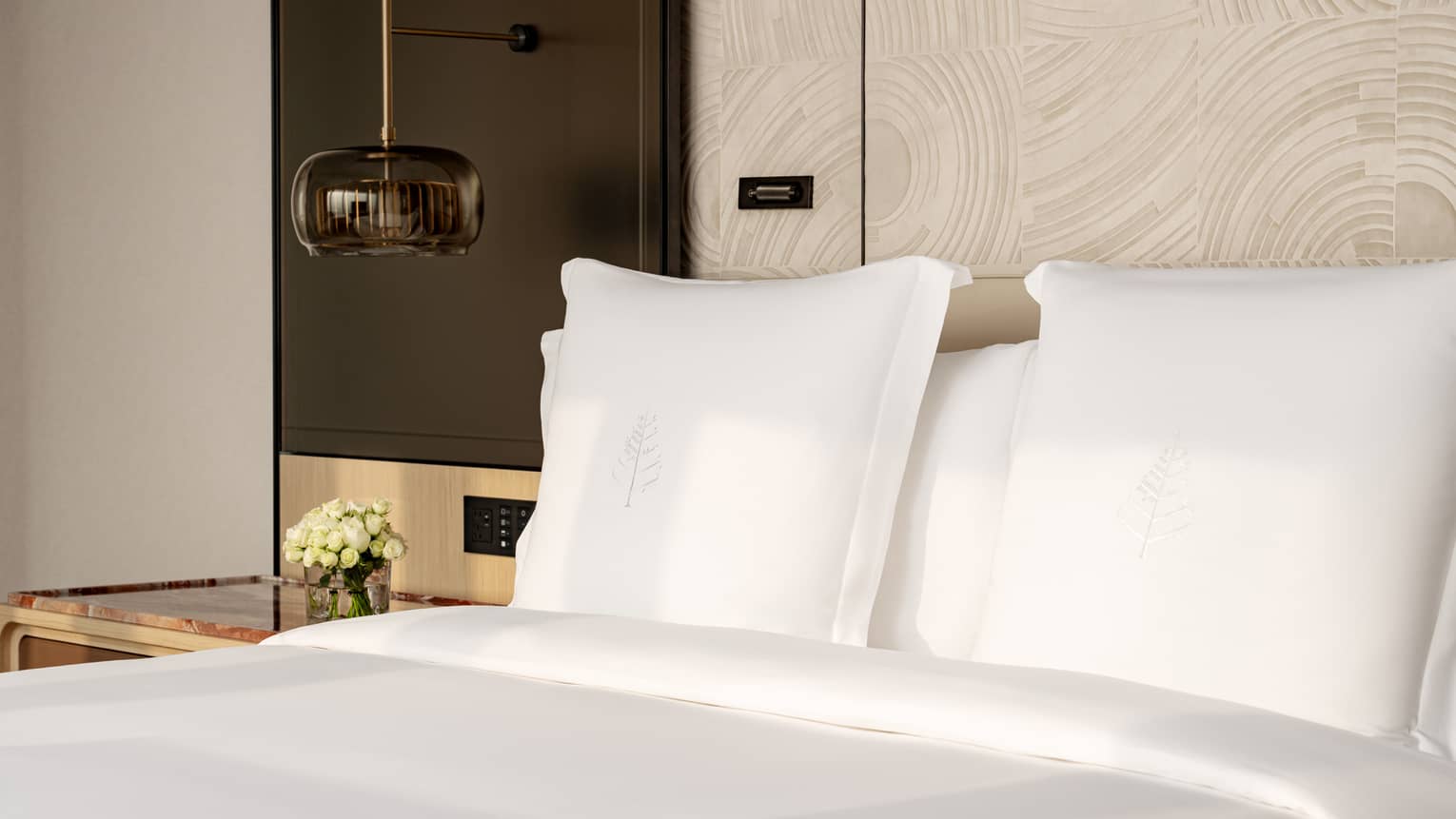 A plush hotel bed, featuring crisp white sheets and shams, set next to pendant light and nightstand with small floral arrangement