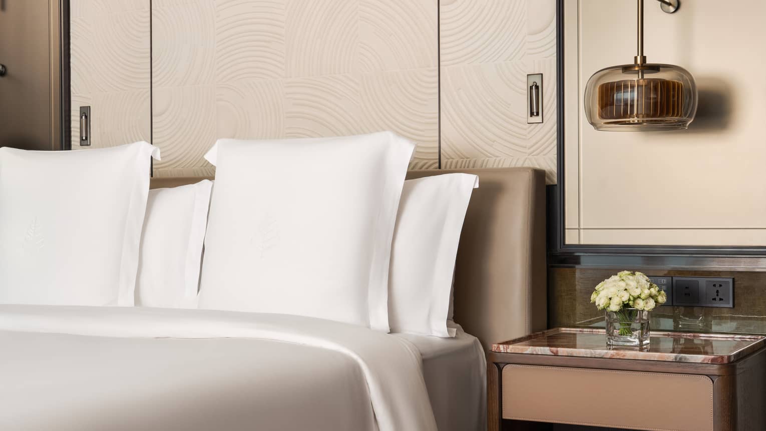 Hotel room bed with crisp white sheets, bedside table holding a small floral arrangement, and a hanging light fixture