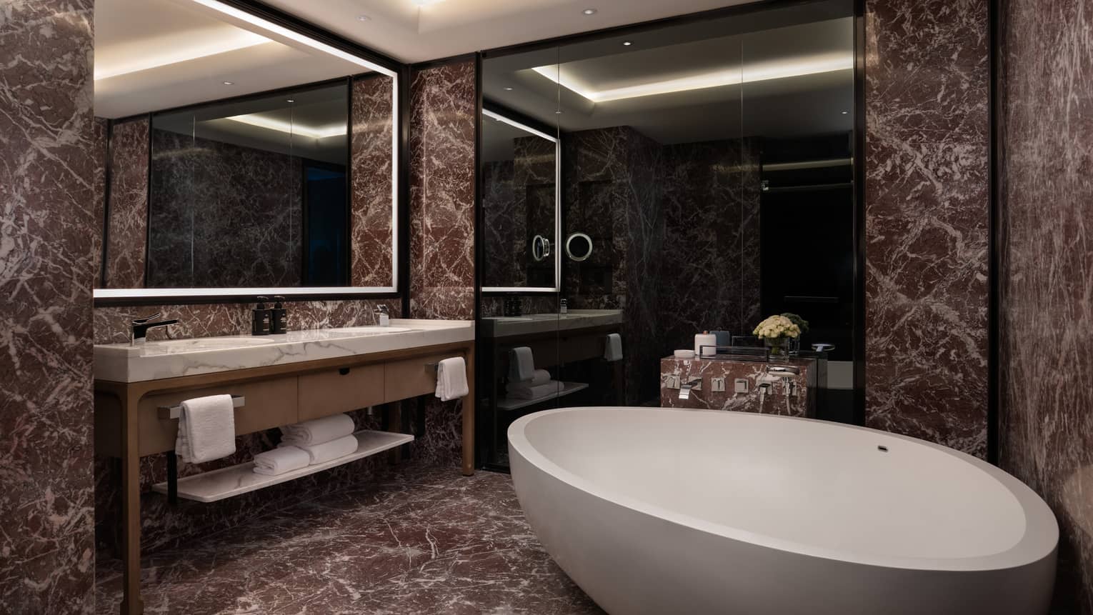 Bathroom with a large standalone tub, dark marble tiles and an illuminated mirror over double vanity