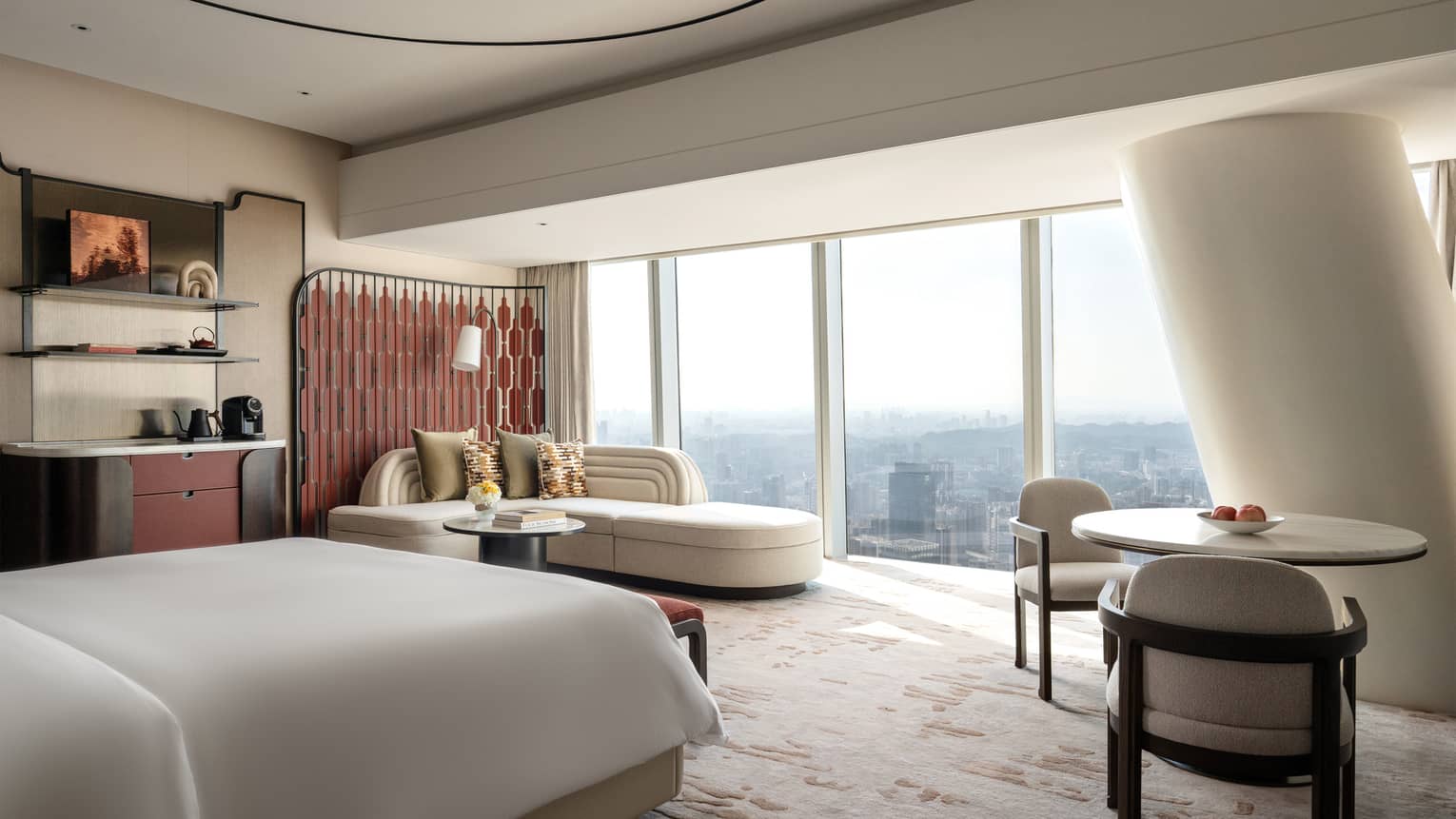 Contemporary hotel room with wall of windows looking out to city, plus a king bed, round table with two chairs and a chaise