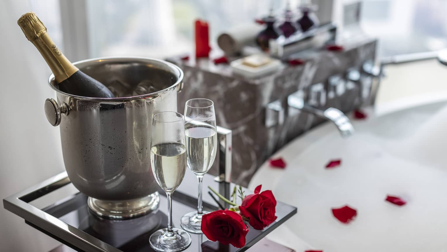 Bottle of champagne in ice bucket and two champagne flutes on tray next to bathtub filled with bubbles and rose petals