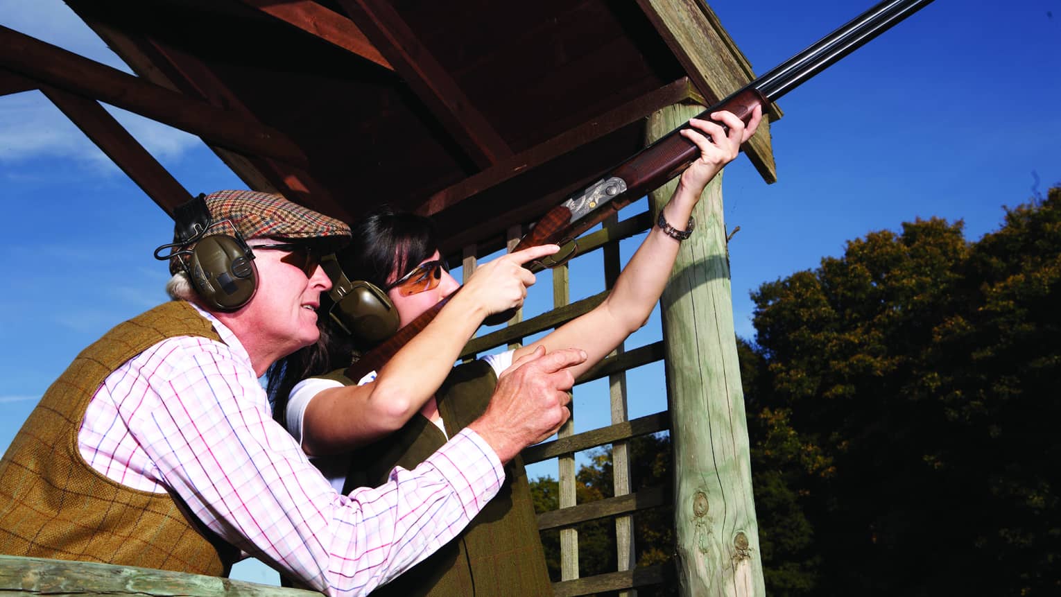 A close-up of two guests wearing tweed vests and shooting ear muffs aiming a rifle towards the clear blue sky.