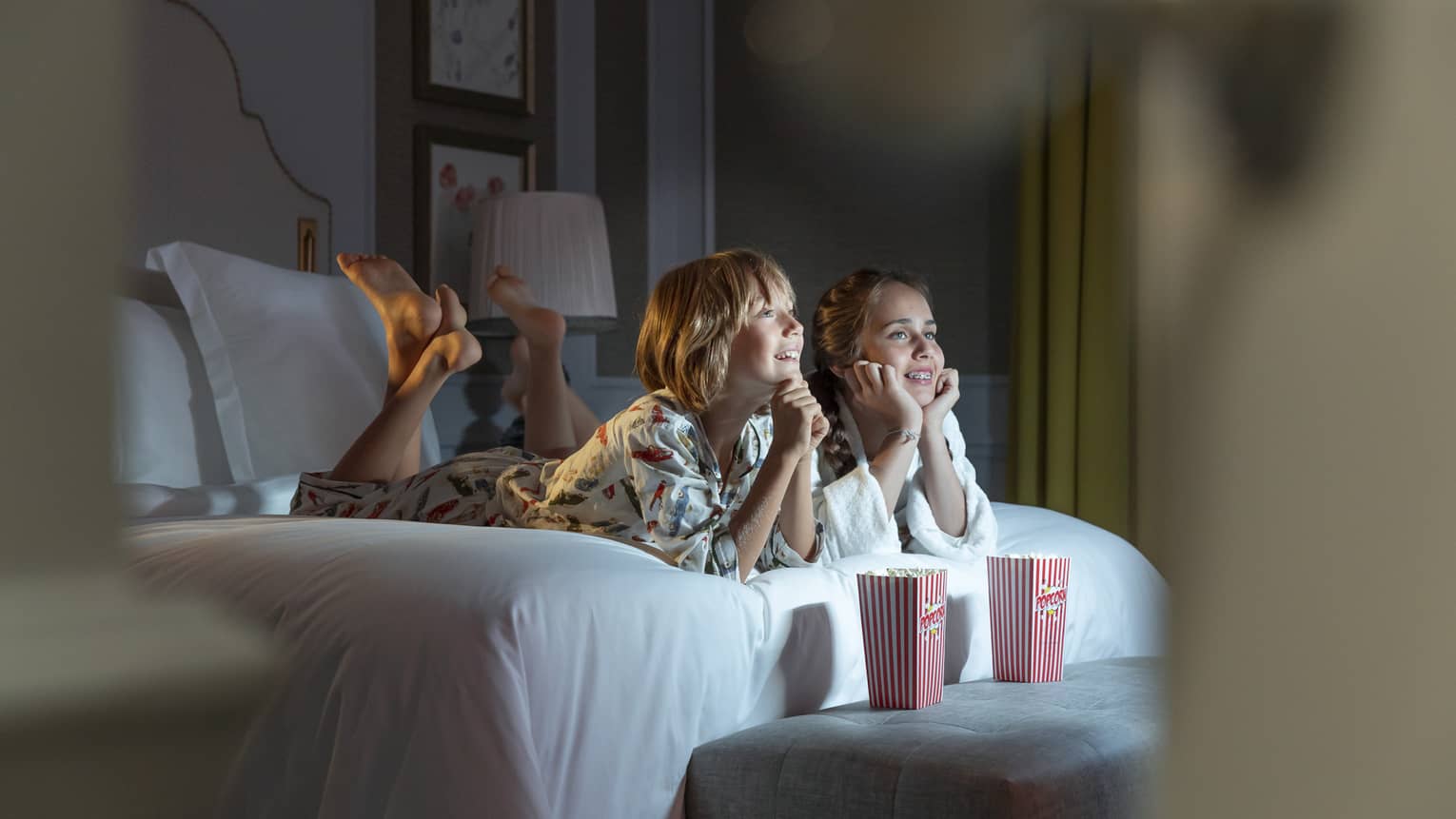 Two children laying on bed and watching tv with popcorn sitting on a bench in front of them.