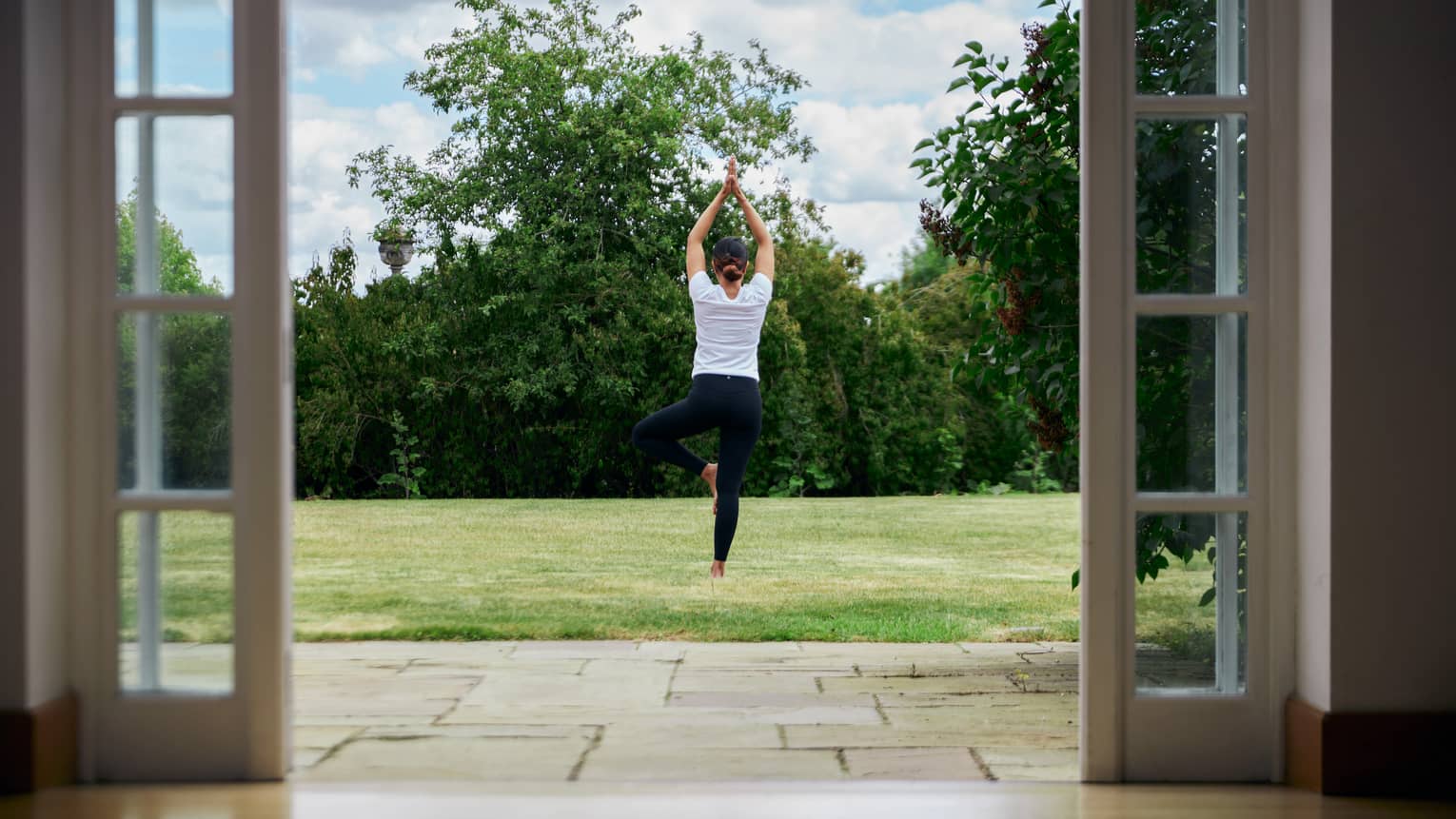 Woman standing in crane yoga pose on green lawn past terrace's French doors