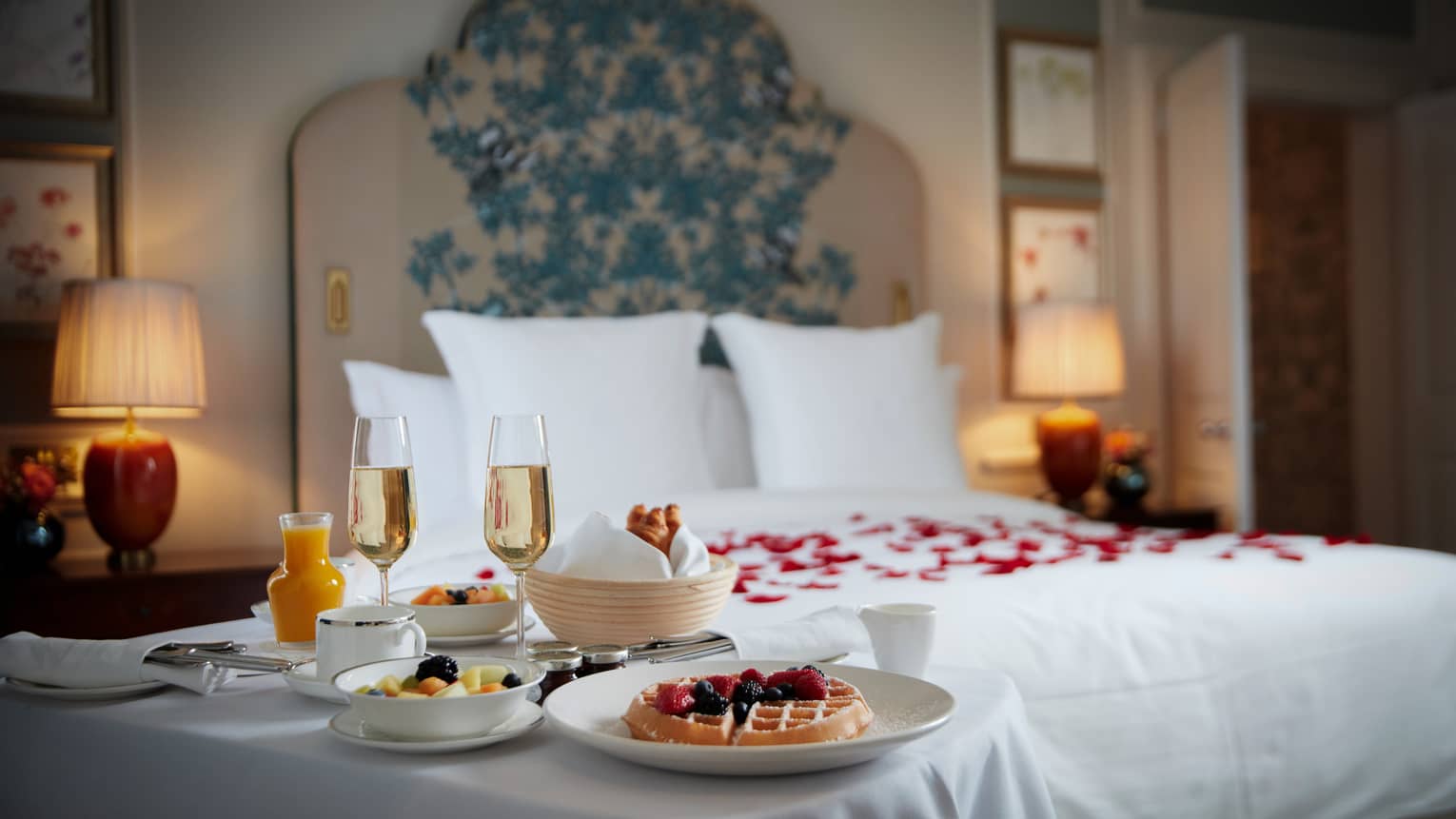 In-Room Dining breakfast set on white clothed table beside white bed with red rose petals