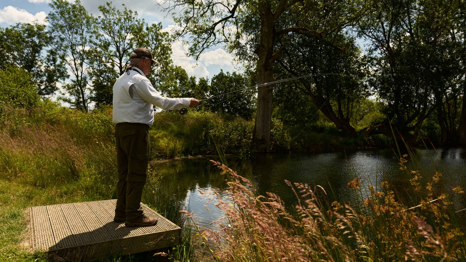 Person in brown cap, white vest and olive green pants standing and fishing off a small pond-side dock