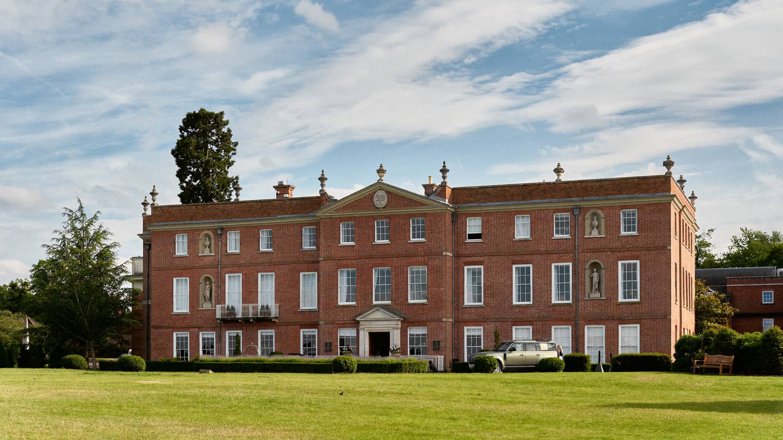 Stately red brick manor house with expansive green lawn under blue sky strewn with clouds